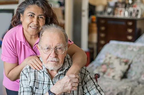 A woman is hugging an older man in his home.