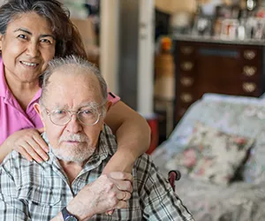 A woman is hugging an older man in his home.